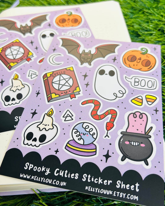 Rebel Romance Spooky Cuties Matte Vinyl Sticker Sheet by Kelly Lou displayed on a notebook with grass background. Includes cute Halloween characters like a bat, ghost, pumpkin, spell book, skull, candle, cauldron, and snake on a lilac background. A waterproof A6 sticker sheet with matte finish—perfect for Halloween crafting, journaling, and spooky gifts.