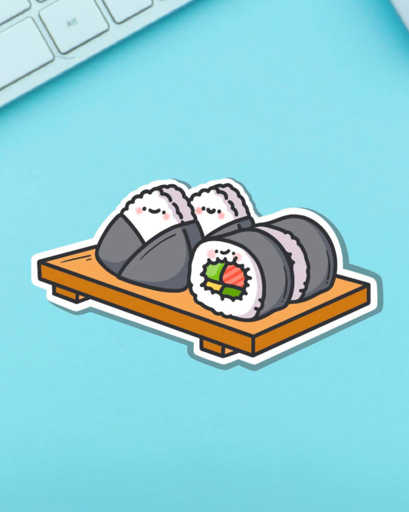 Glossy sushi vinyl sticker featuring happy-faced sushi rolls and onigiri, laid on a wooden tray. Styled on a bright turquoise surface next to a keyboard. From Rebel Romance, a small UK biz selling fun and alternative fashion accessories.