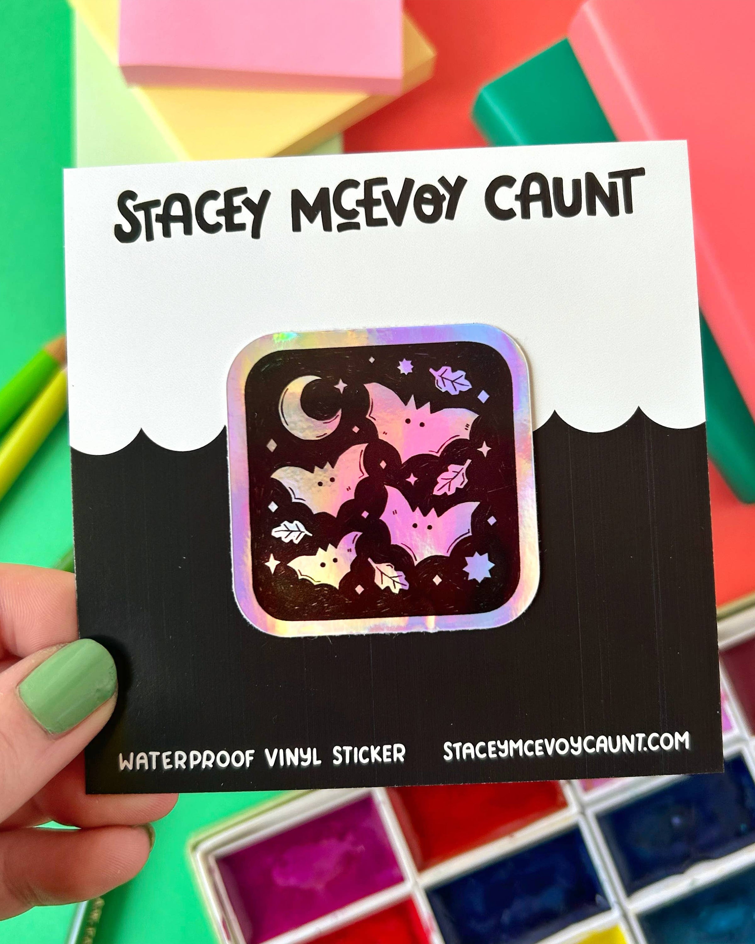 Square holographic bat sticker displayed on a black and white branded backing card from Stacey McEvoy Caunt. The sticker features a spooky-cute bat pattern with stars, moons, and leaves on a shiny rainbow finish. Held in front of colourful stationery and a watercolour palette. Sold by Rebel Romance, a small UK business celebrating alternative millennial style.