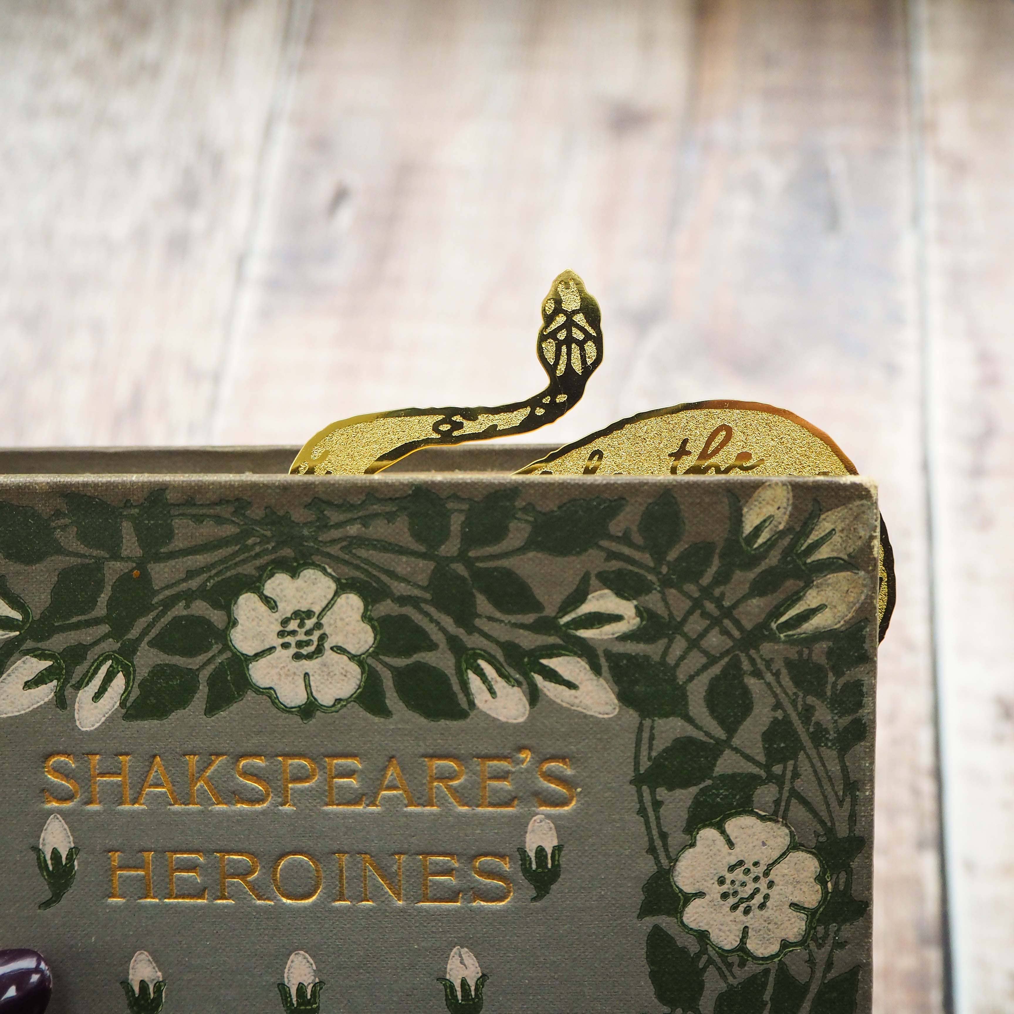 A brass bookmark shaped like a coiled serpent, delicately etched with the famous Lady Macbeth quote: “Look like the innocent flower, But be the serpent under ‘t.” The bookmark is placed inside an open book on a wooden surface, with the title “Lady Macbeth” visible on the page. The snake’s head is designed to peek out from the top of the book when closed, adding a dramatic and literary flair.