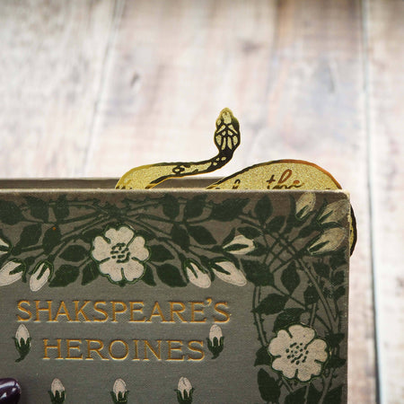 A brass bookmark shaped like a coiled serpent, delicately etched with the famous Lady Macbeth quote: “Look like the innocent flower, But be the serpent under ‘t.” The bookmark is placed inside an open book on a wooden surface, with the title “Lady Macbeth” visible on the page. The snake’s head is designed to peek out from the top of the book when closed, adding a dramatic and literary flair.
