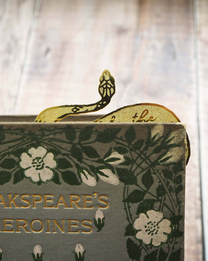 A brass bookmark shaped like a coiled serpent, delicately etched with the famous Lady Macbeth quote: “Look like the innocent flower, But be the serpent under ‘t.” The bookmark is placed inside an open book on a wooden surface, with the title “Lady Macbeth” visible on the page. The snake’s head is designed to peek out from the top of the book when closed, adding a dramatic and literary flair.