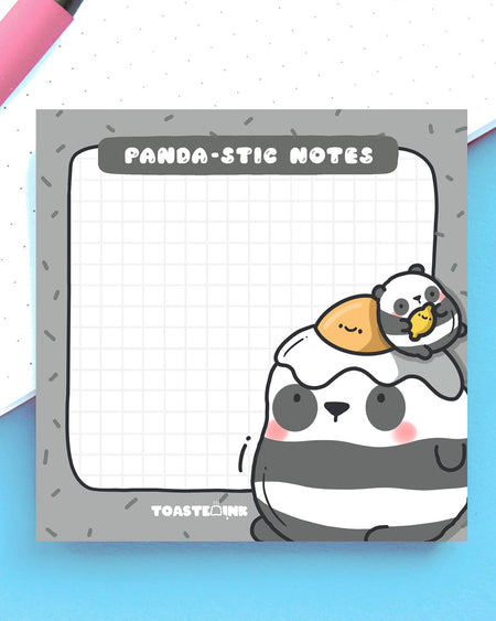 Adorable panda-themed sticky notes by Toastedink, shown flat on a desk with a pink pen. Grid design with "Panda-stic Notes" pun, stocked by Rebel Romance – the home of weird, wonderful, and wildly cute stationery.