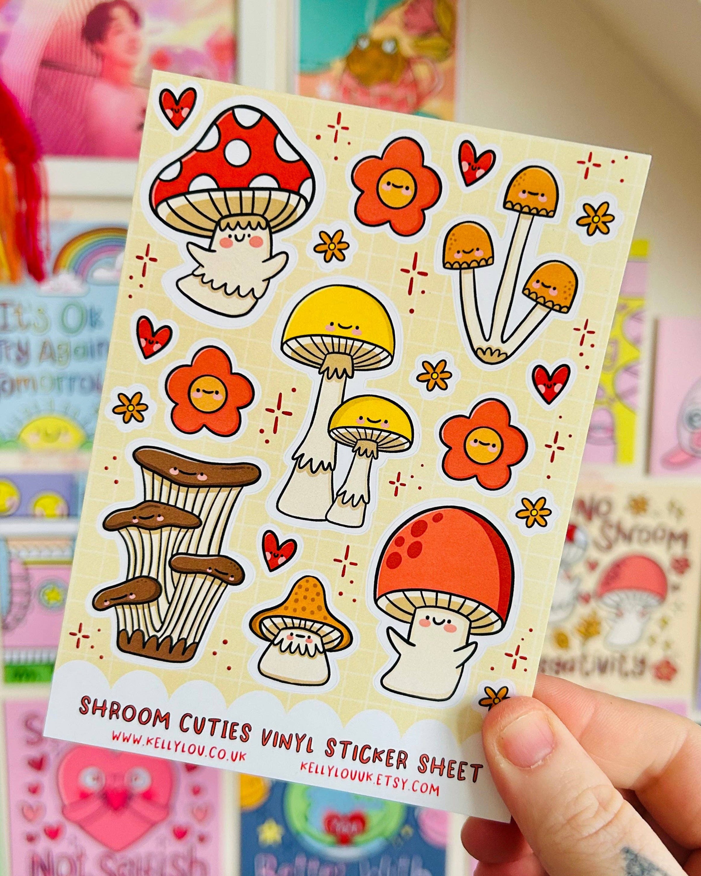 Hand holding the Shroom Cuties Vinyl Sticker Sheet by Kelly Lou, sold by Rebel Romance. The sheet showcases smiling cartoon mushrooms in vibrant red, yellow, and orange with floral and heart designs on a beige background. Ideal for adding a whimsical touch to notebooks or gifts. Colourful art prints and cards can be seen blurred in the background.
