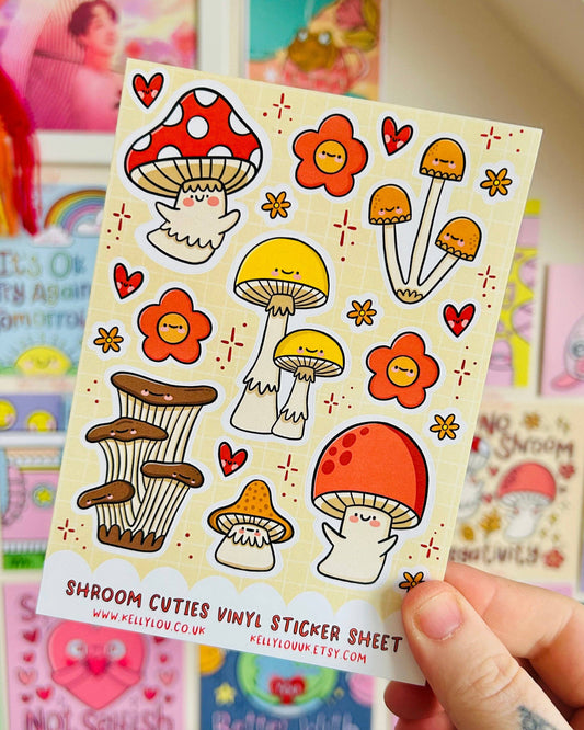 Hand holding the Shroom Cuties Vinyl Sticker Sheet by Kelly Lou, sold by Rebel Romance. The sheet showcases smiling cartoon mushrooms in vibrant red, yellow, and orange with floral and heart designs on a beige background. Ideal for adding a whimsical touch to notebooks or gifts. Colourful art prints and cards can be seen blurred in the background.