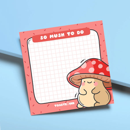 A square sticky note pad with a red border and grid paper design, featuring a cute, blushing mushroom character with a red and white spotted cap in the bottom right corner. The top header reads “So Mush To Do” in bold white text. A fun and functional memo pad for everyday reminders.