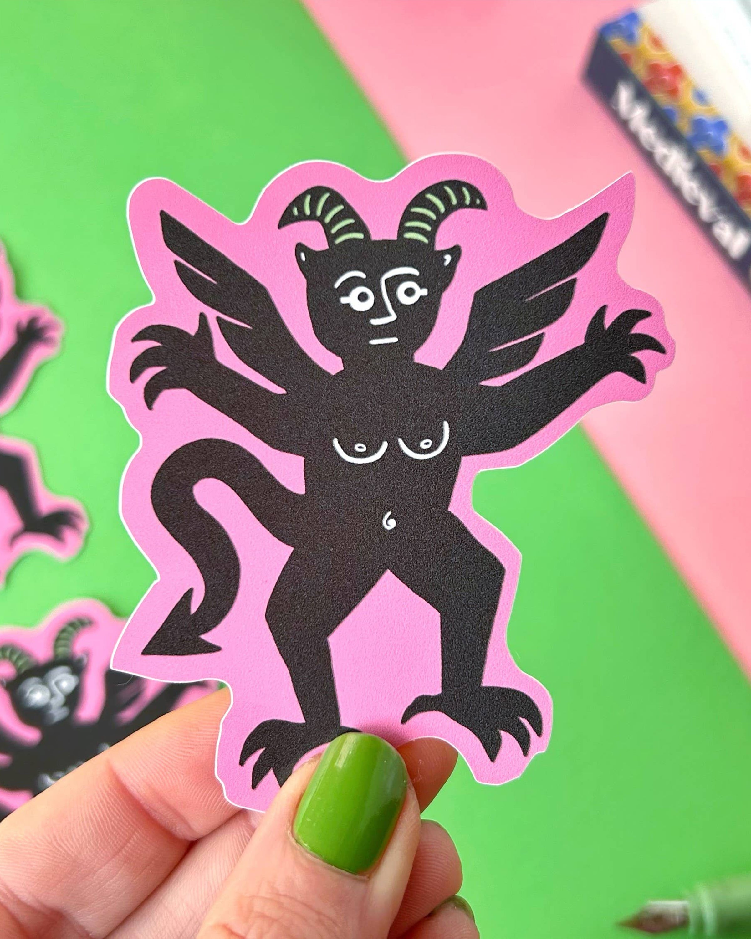 Close-up of a hand with green nail polish holding the Pink Devil Sticker by Stacey McEvoy Caunt. The sticker shows a six-armed black devil with wings and horns, printed on a bright pink background. Rebel Romance stocks this alternative vinyl sticker. Colourful books and other devil stickers are visible in the background.
