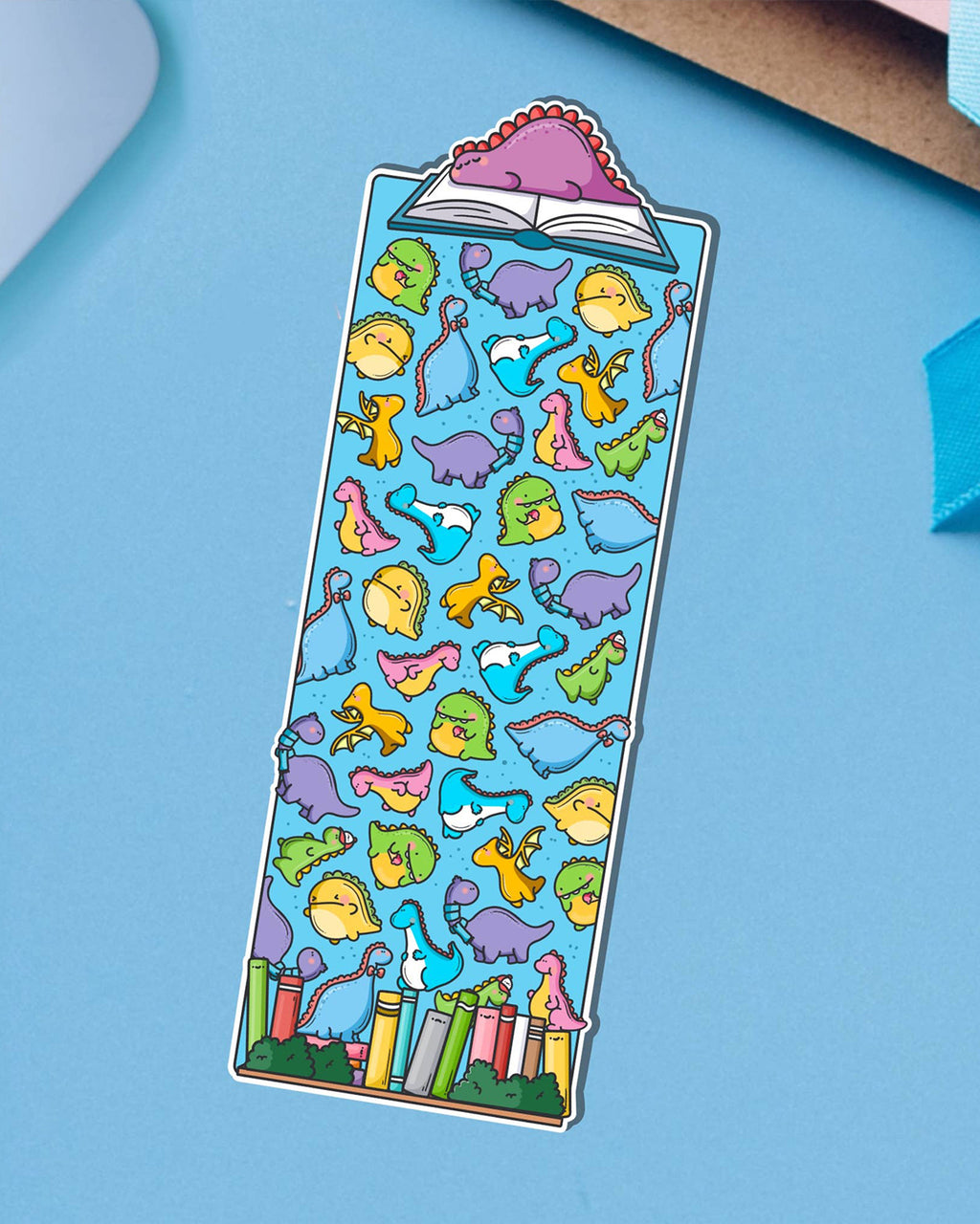 Rebel Romance dinosaur bookmark by Toastedink, printed with a cheerful herd of multicoloured dinos and books at the base. Topped with a pink dinosaur lounging on a book. Set on a bright blue background.