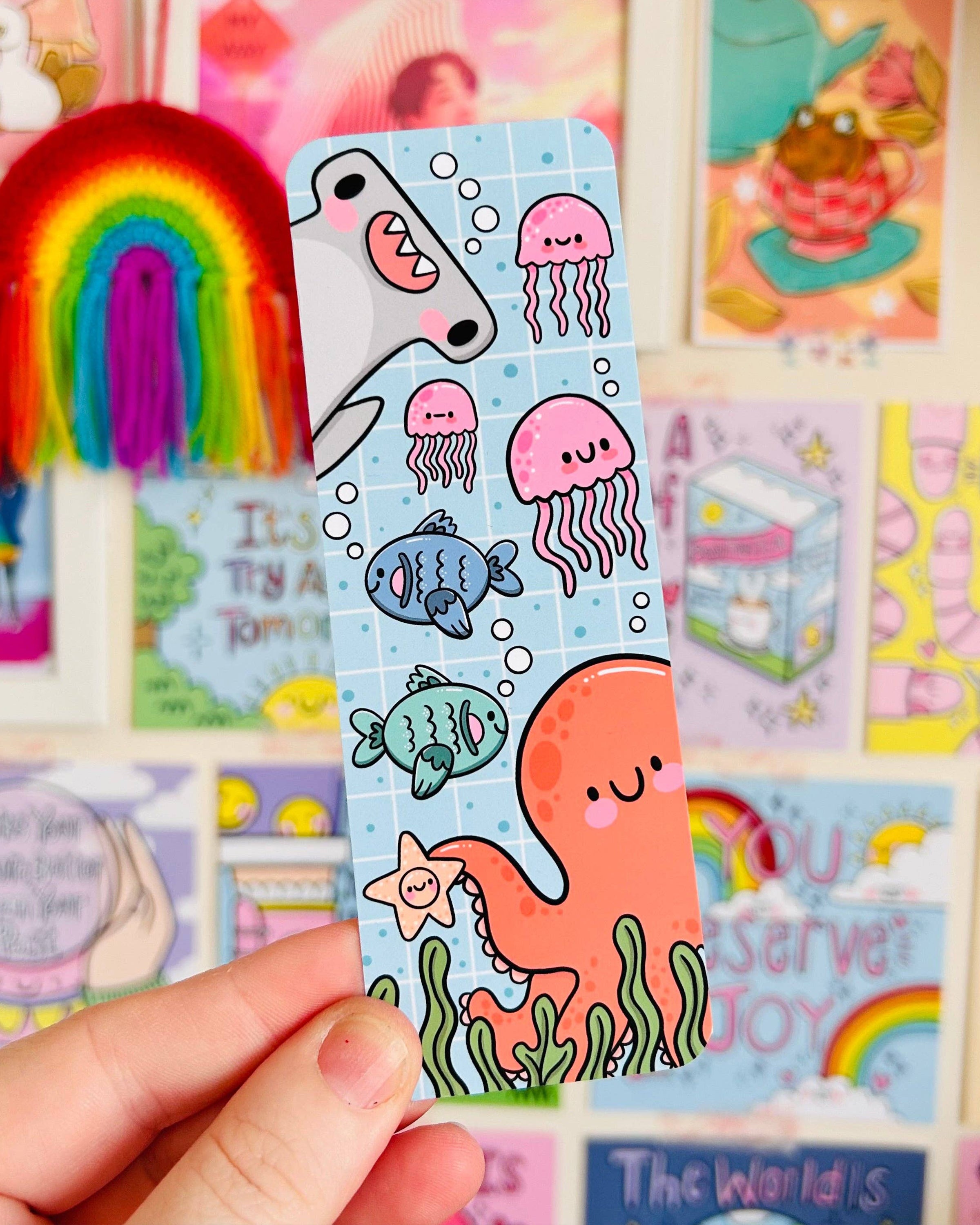 Rebel Romance Under The Sea bookmark by Kelly Lou held up against a colourful wall of motivational prints and rainbows. Features a smiling orange octopus, pink jellyfish, cartoon fish, and a cheerful shark. Made from velvet-laminated card with a glossy finish, perfect for ocean lovers and whimsical readers. A fun gift for bookworms and stationery fans.