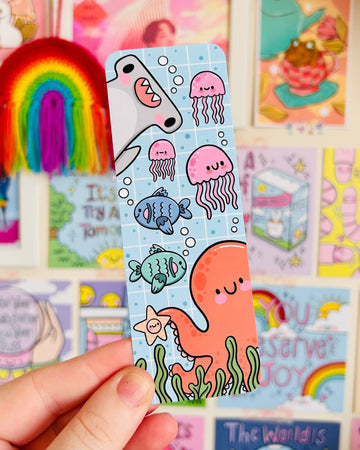 Rebel Romance Under The Sea bookmark by Kelly Lou held up against a colourful wall of motivational prints and rainbows. Features a smiling orange octopus, pink jellyfish, cartoon fish, and a cheerful shark. Made from velvet-laminated card with a glossy finish, perfect for ocean lovers and whimsical readers. A fun gift for bookworms and stationery fans.