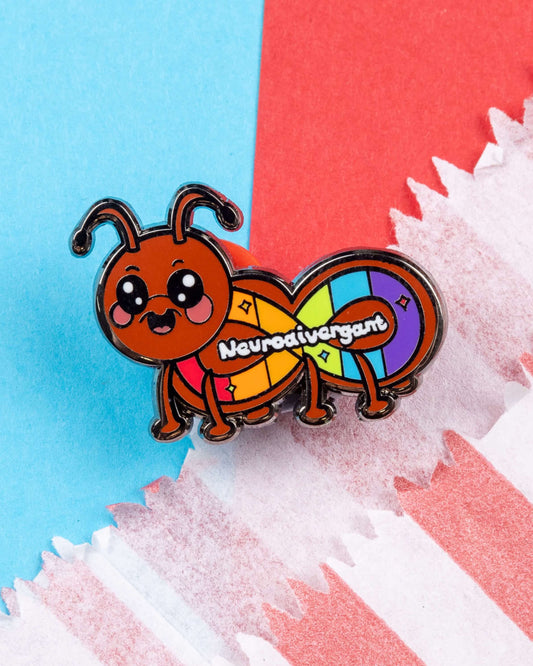 Close-up of the Rebel Romance Neurodivergant enamel pin designed by Innabox, shaped like a happy red ant with rainbow body segments and the word "Neurodivergant" across it. Displayed on a red, white, and blue paper background. A colourful, quirky pin that celebrates neurodiversity with pride and humour.