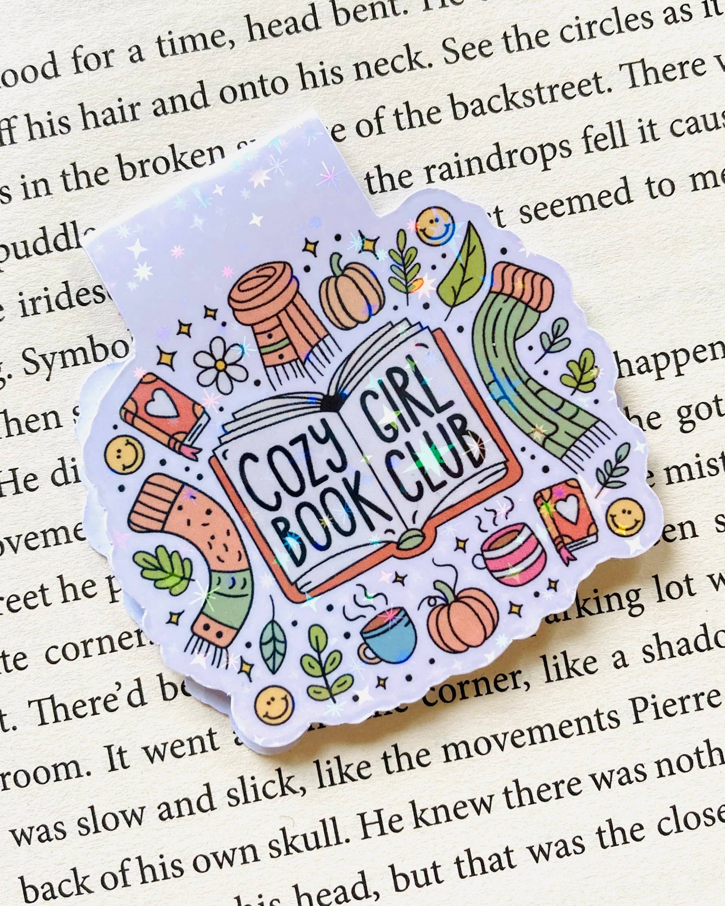 Close‑up of the Rebel Romance “Cozy Girl Book Club” magnetic bookmark by The Stickityboo Co clipped onto a book page. The design shows colourful scarves, coffee cups, pumpkins and plants with a whimsical, glittery finish, perfect for cosy reading sessions.