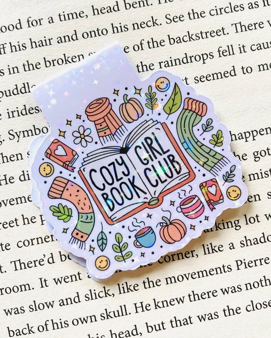 Close‑up of the Rebel Romance “Cozy Girl Book Club” magnetic bookmark by The Stickityboo Co clipped onto a book page. The design shows colourful scarves, coffee cups, pumpkins and plants with a whimsical, glittery finish, perfect for cosy reading sessions.
