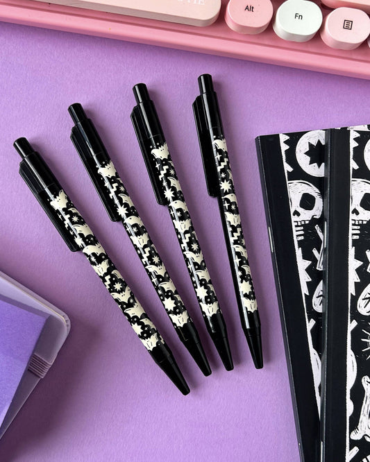 Flat lay of four Rebel Romance Bat Pattern Pens arranged diagonally on a purple background, next to matching gothic black-and-white notebooks and a pastel keyboard.