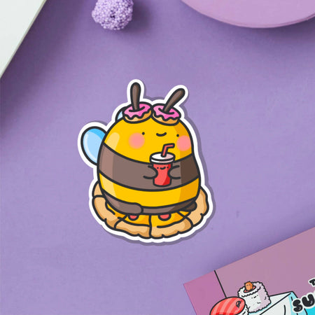 Cute cartoon-style vinyl sticker of a bumblebee with pink doughnut antennae, holding a red drink and sitting on a pie. Ideal for laptops, bottles, and journals.