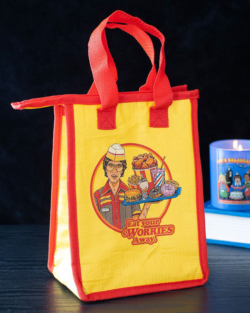 Lifestyle shot of Rebel Romance’s Steven Rhodes “Eat Your Worries Away” insulated lunch bag placed on a black surface with a candle in the background. Bold yellow and red design with quirky retro food artwork, perfect for storing snacks, leftovers, or daily lunches with humour.