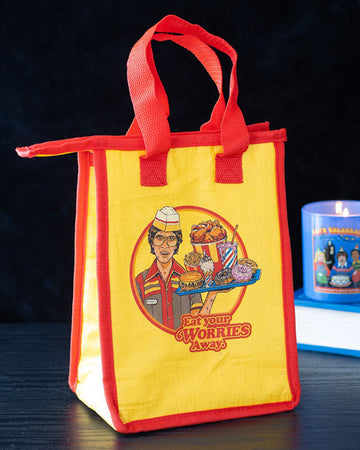 Lifestyle shot of Rebel Romance’s Steven Rhodes “Eat Your Worries Away” insulated lunch bag placed on a black surface with a candle in the background. Bold yellow and red design with quirky retro food artwork, perfect for storing snacks, leftovers, or daily lunches with humour.