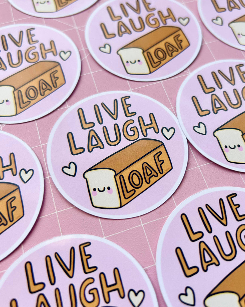 Multiple Rebel Romance “Live Laugh Loaf” glossy stickers by Kelly Lou laid out on a pink grid background. Each circular sticker shows a smiling loaf of bread with hearts, celebrating alternative, playful aesthetics loved by millennials.