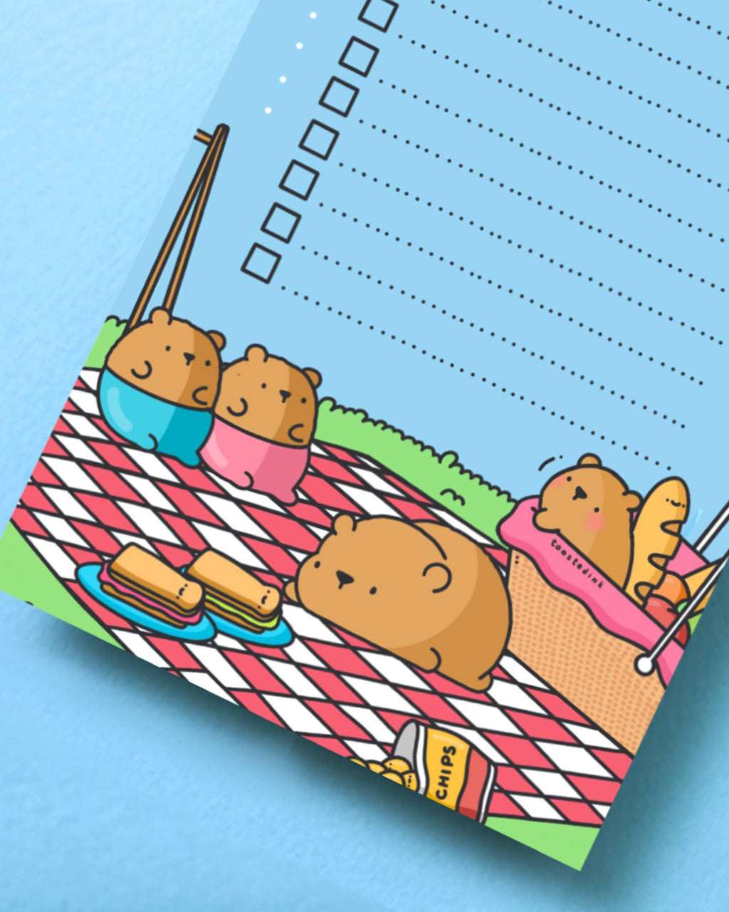 Close-up of the bottom half of the Toastedink bear to-do list pad showing bears having a picnic on a red chequered blanket with sandwiches and crisps, against a blue sky background.