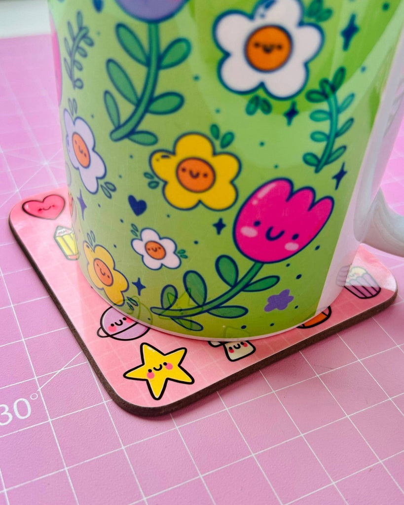 A close-up of the Appreciate The Little Things drinks coaster by Kelly Lou from Rebel Romance, a small UK brand celebrating alternative style. The coaster features cute illustrations including a star, mushrooms and smiley faces on a pink background, with a colourful floral mug resting on top.