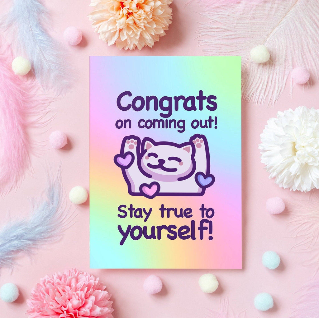 Coming out greeting card by Mona Mauve for Rebel Romance displayed on a pink background with soft feathers, pom poms, and flowers. Features a pastel rainbow design with a smiling cartoon cat and uplifting text: “Congrats on coming out! Stay true to yourself!” A heartfelt and inclusive LGBTQIA+ celebration card.