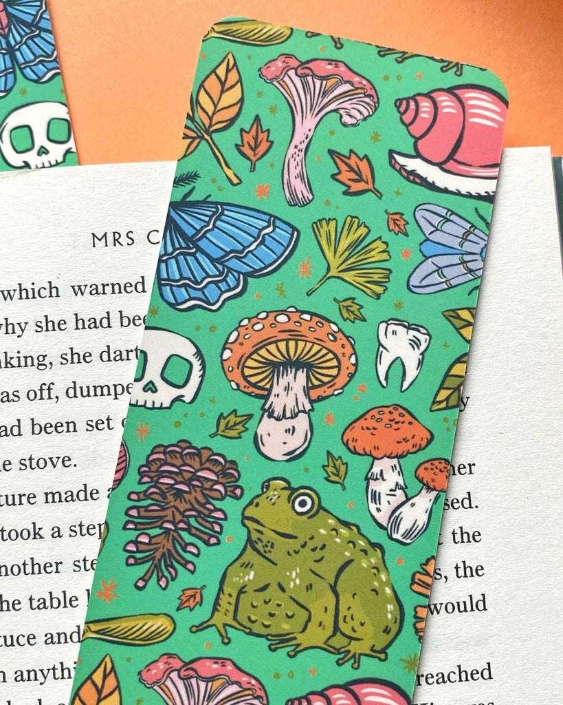 A green bookmark with a pattern of mushrooms, leaves, pinecones, and a large toad illustration. The background features an open book on an orange surface.