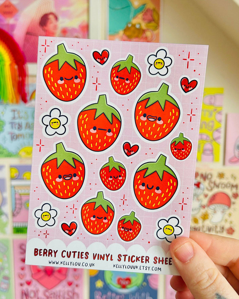 Rebel Romance Berry Cuties Vinyl Sticker Sheet by Kelly Lou held up in front of a colourful wall of prints. Includes cute cartoon strawberries with happy faces, floral accents, and hearts on a pastel pink grid background. Waterproof and glossy A6 sheet, great for journaling, scrapbooking, or gifting to someone who loves fruit-themed stationery and fun designs.