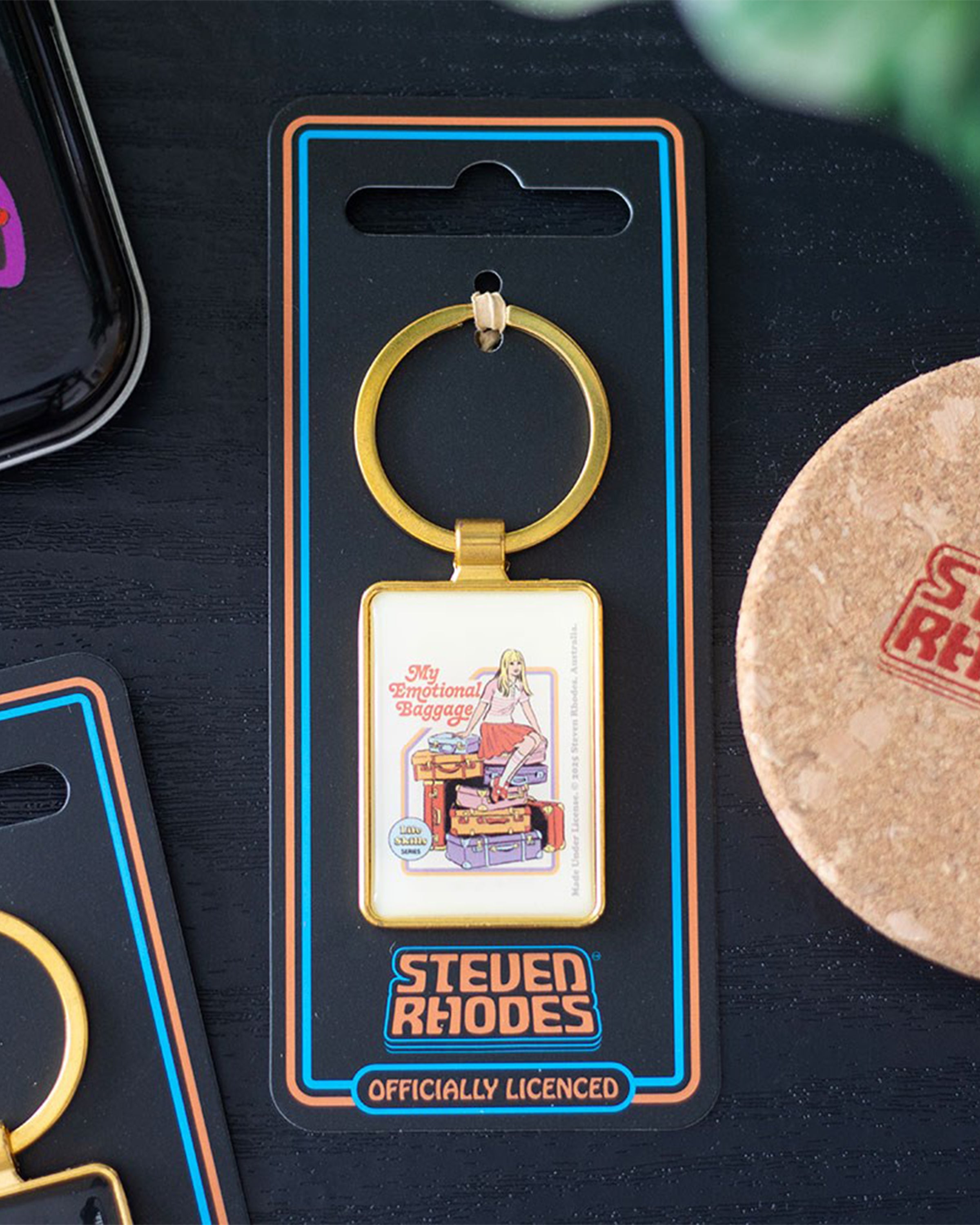 Rebel Romance enamel keyring on Steven Rhodes branded packaging, featuring the design “My Emotional Baggage.” The gold retro-inspired accessory shows a girl sitting on colourful luggage, making it a fun, alternative and quirky gift idea with bold humour and style.
