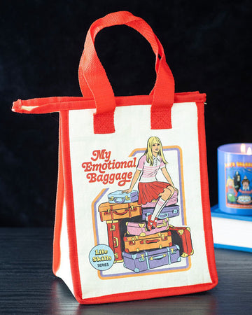 Rebel Romance Steven Rhodes insulated lunch bag "My Emotional Baggage" displayed on a black table beside a retro candle. Cream with red trim and handles, featuring fun vintage-style illustration. A quirky eco-friendly lunch tote made from recycled plastic.