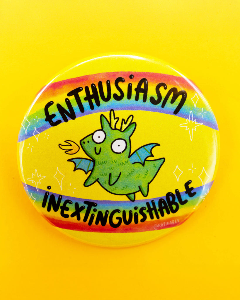 Bright yellow background showcasing a round pin badge by Katie Abey for Rebel Romance. The badge features a cheerful green dragon with wings, fire breath, and rainbow accents. The text reads “Enthusiasm Inextinguishable” in bold black lettering. A bold and magical design from a UK small business celebrating weird and wonderful aesthetics.