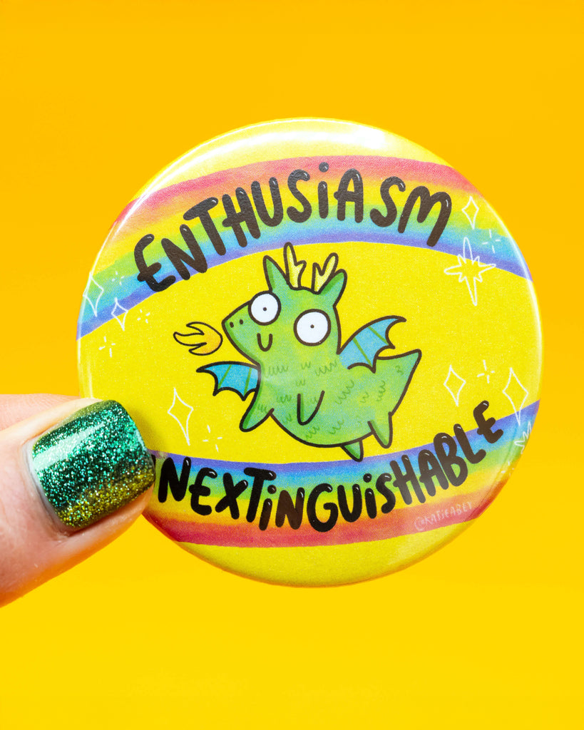 Hand holding a colourful round badge designed by Katie Abey for Rebel Romance, featuring a cartoon green dragon with wings and flames, surrounded by the words “Enthusiasm Inextinguishable” across rainbow stripes on a yellow background. A playful gift from a UK-based alternative fashion and lifestyle brand.