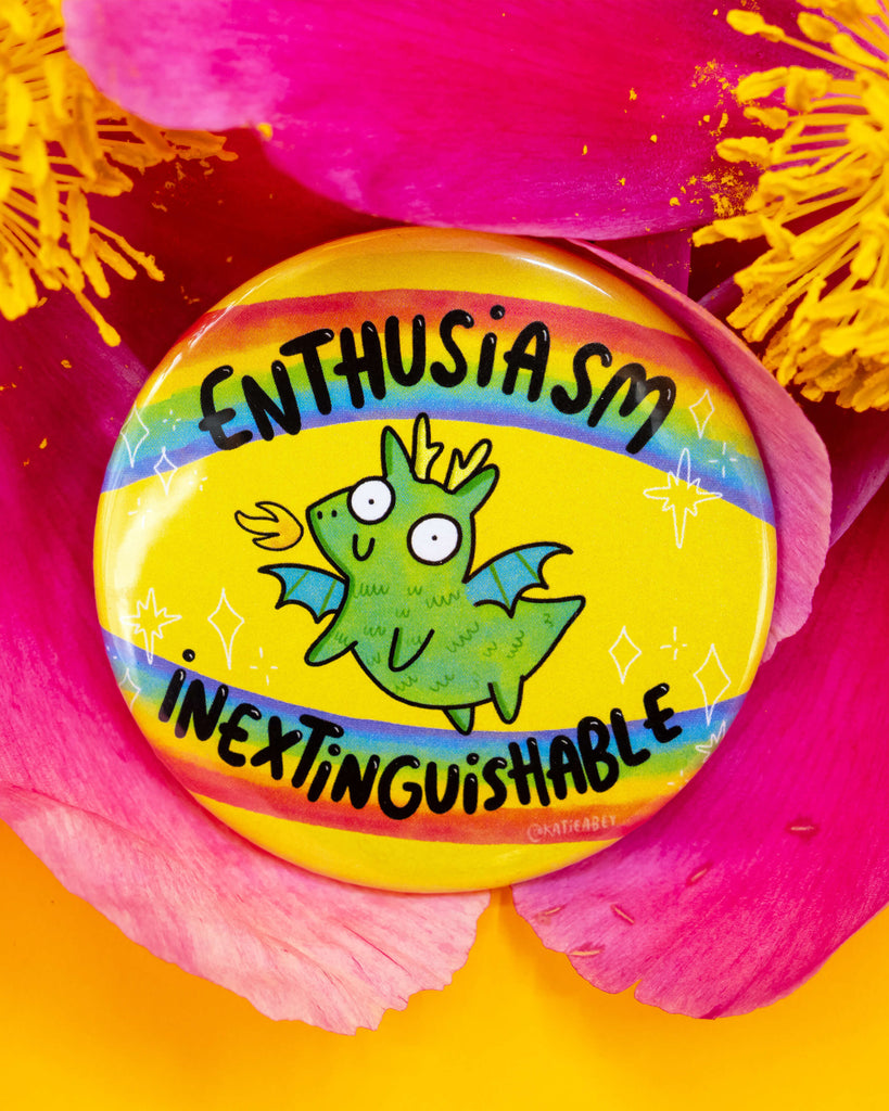 Close-up of a bright yellow badge by Katie Abey for Rebel Romance featuring a quirky green dragon breathing fire with the phrase “Enthusiasm Inextinguishable” in bold black letters on rainbow stripes. The badge is placed inside vibrant pink and yellow flower petals. A fun and colourful accessory by a small UK business celebrating alternative style.