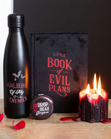 A gothic gift flat lay featuring Rebel Romance’s Little Book of Evil Plans velvet notebook, a black bottle reading "The Blood of My Enemies", a compact mirror that says "Drop Dead Gorgeous", and a black and red dripping candle, styled on a wooden surface with rose petals.