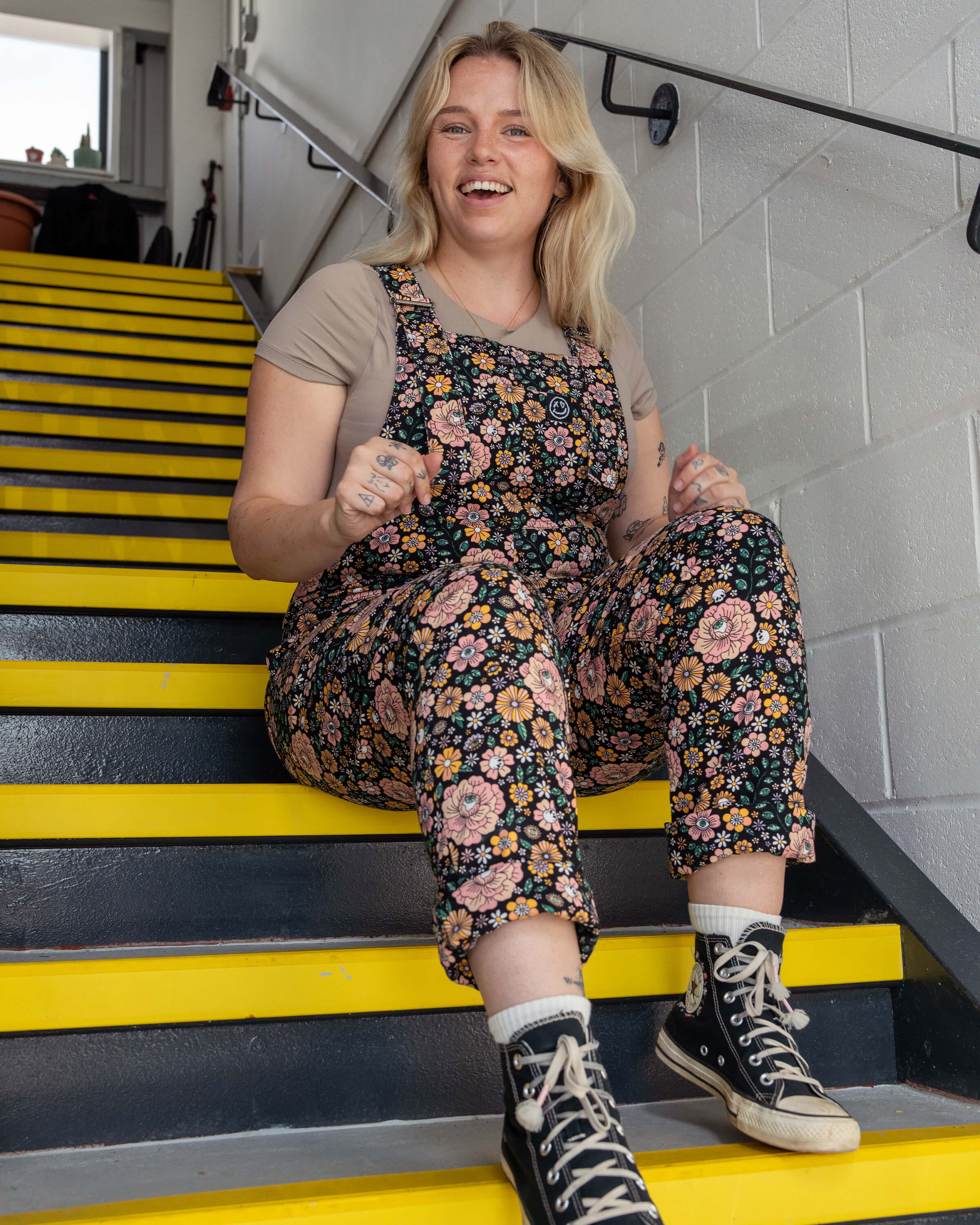 Harleigh sitting on yellow stairs wearing Rebel Romance Eye Spy Floral Stretch Dungarees in floral and skull print with a beige tee.