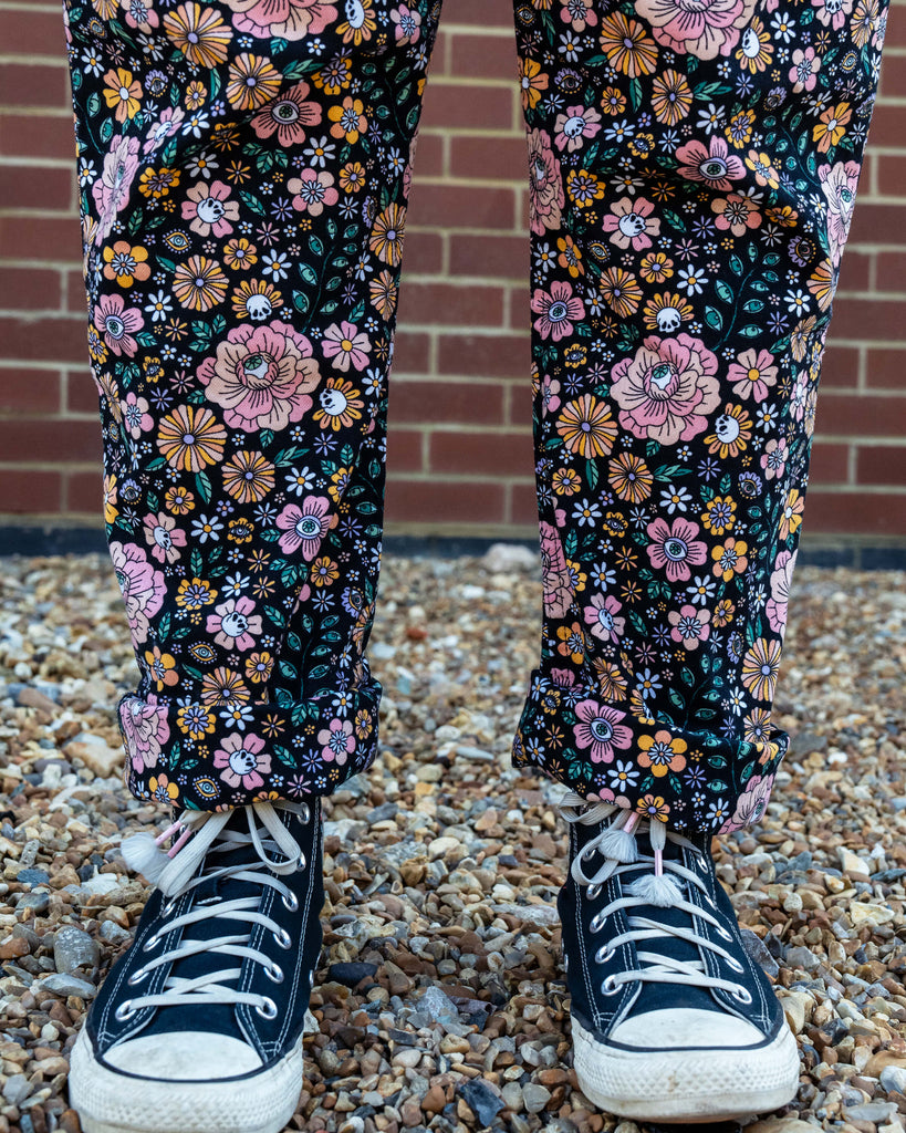 Close-up of Rebel Romance Eye Spy Floral Stretch Dungarees in black corduroy with pastel pink, orange, and yellow flower print, featuring hidden skull and eye motifs, styled with black Converse trainers.