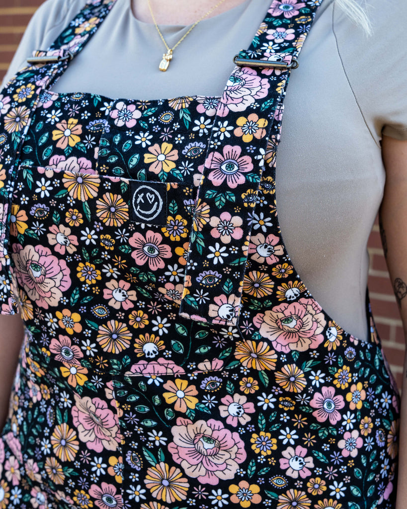 Front bib detail of Rebel Romance Eye Spy Floral Stretch Dungarees showing the floral, skull, and eye design in pink, orange, and green on a black background, modelled with a beige top.
Back view of Rebel Romance Eye Spy Floral Stretch Dungarees on Harleigh, showing the all-over floral and skull print design against a brick wall backdrop.