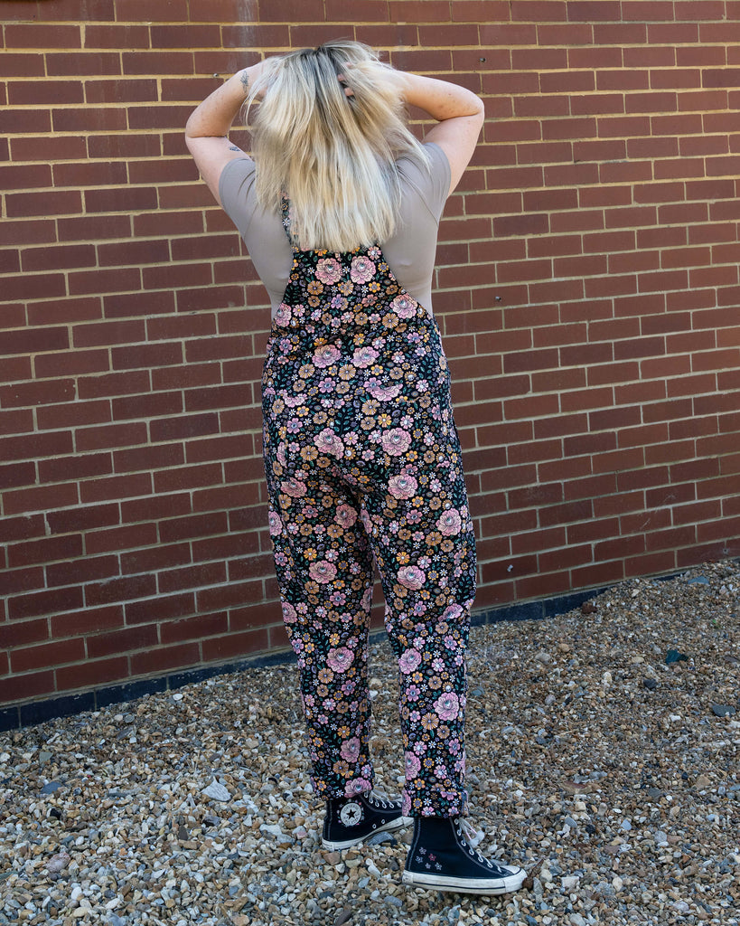 Back view of Rebel Romance Eye Spy Floral Stretch Dungarees on Harleigh, showing the all-over floral and skull print design against a brick wall backdrop.