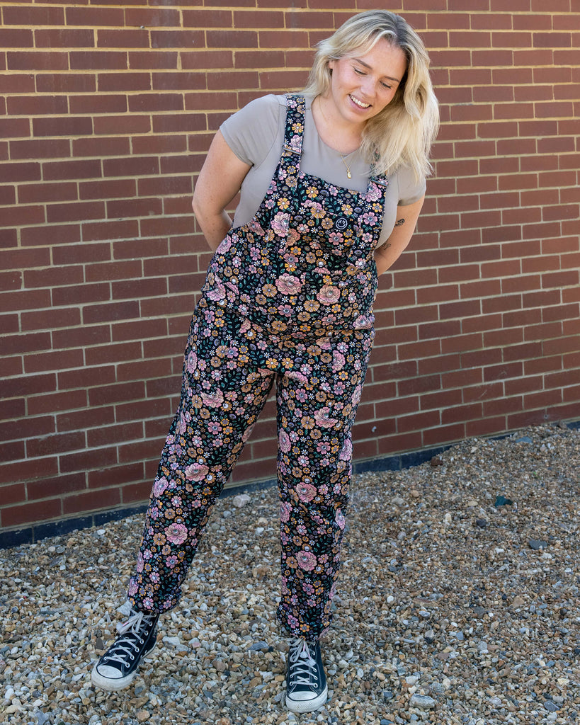 Woman wearing floral overalls standing against a brick wall.
