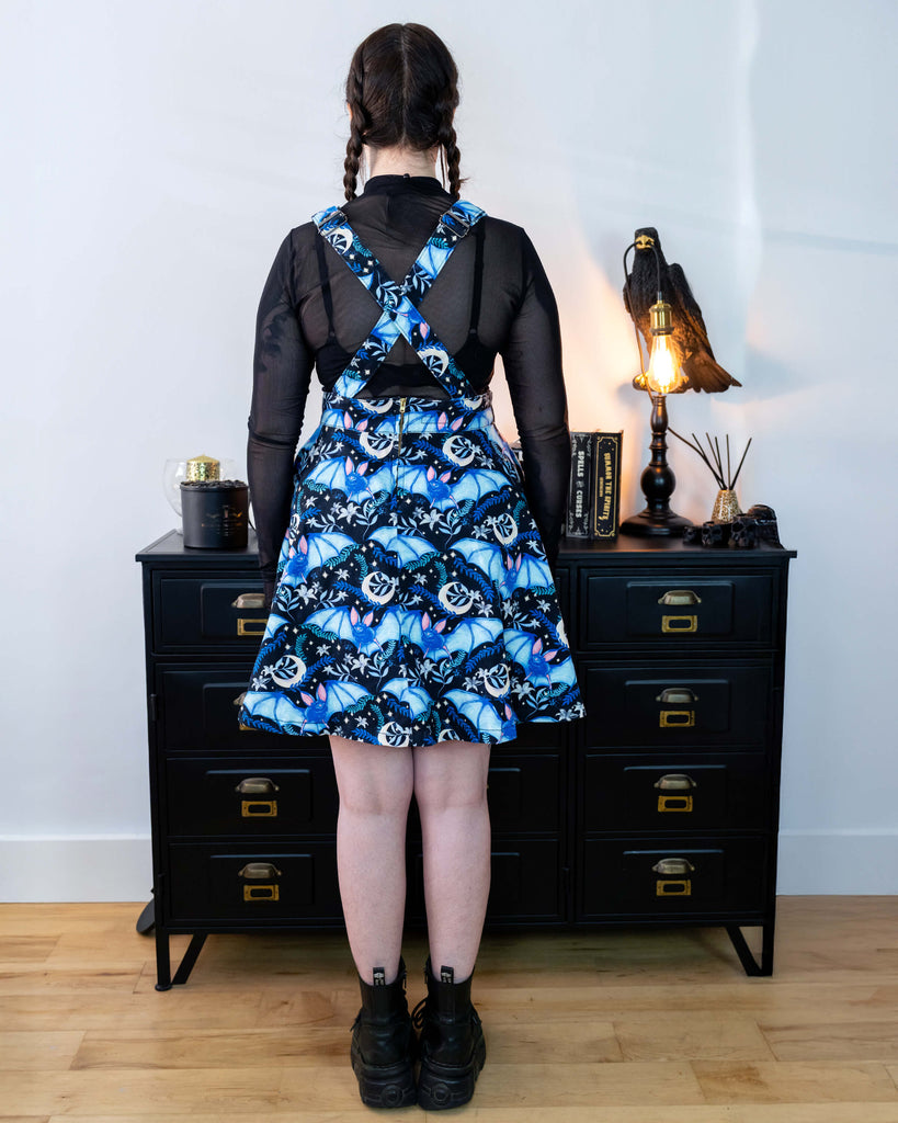 Alice is stood in front of a black dresser with creepy books and a crow lamp she is wearing a night flight flared pinafore and looks like Wednesday Addams. She has a black mesh top underneath. Alice has her back to the camera and you can see the cross over of the straps on the back.