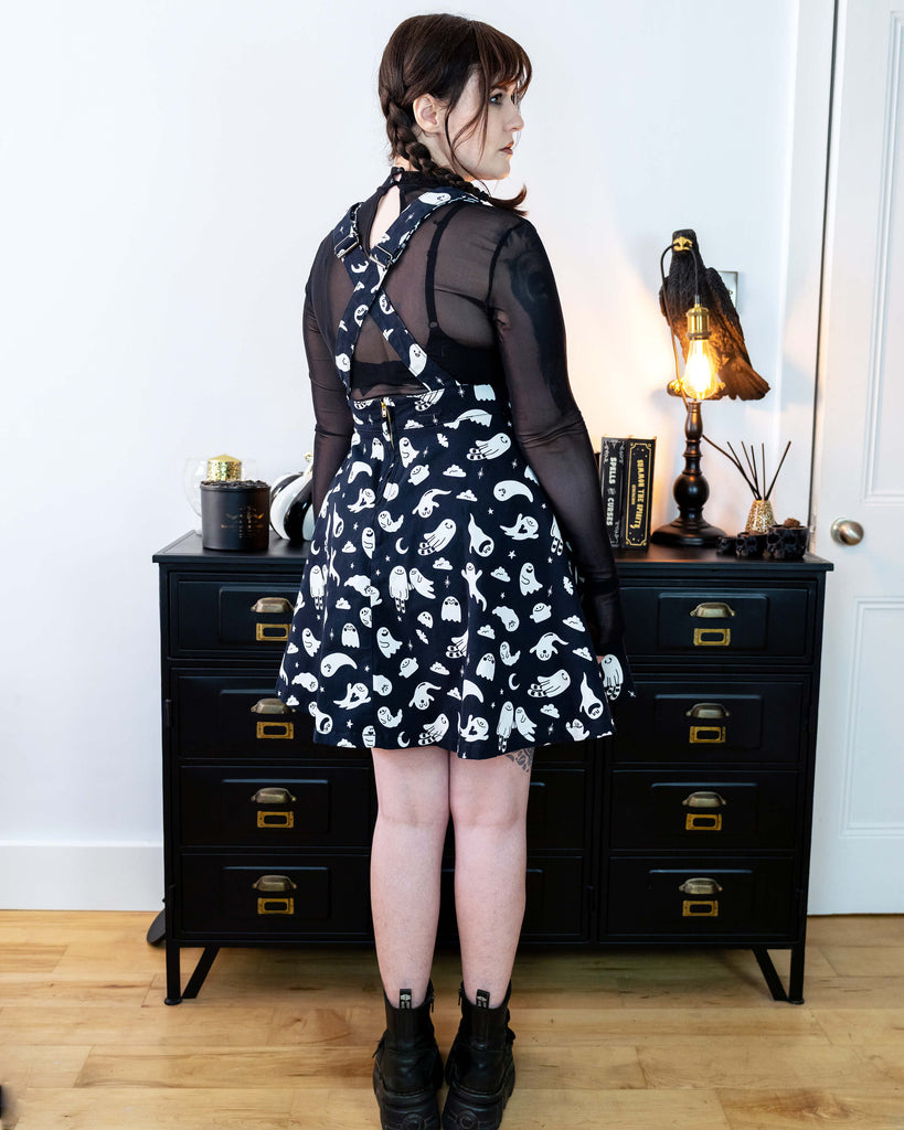 Femme model with brown plaited hair facing away from camera wearing a black flared pinafore dress with a ghost pattern and a mesh black top underneath in a gothic styled room with a crow lamp and black drawers.