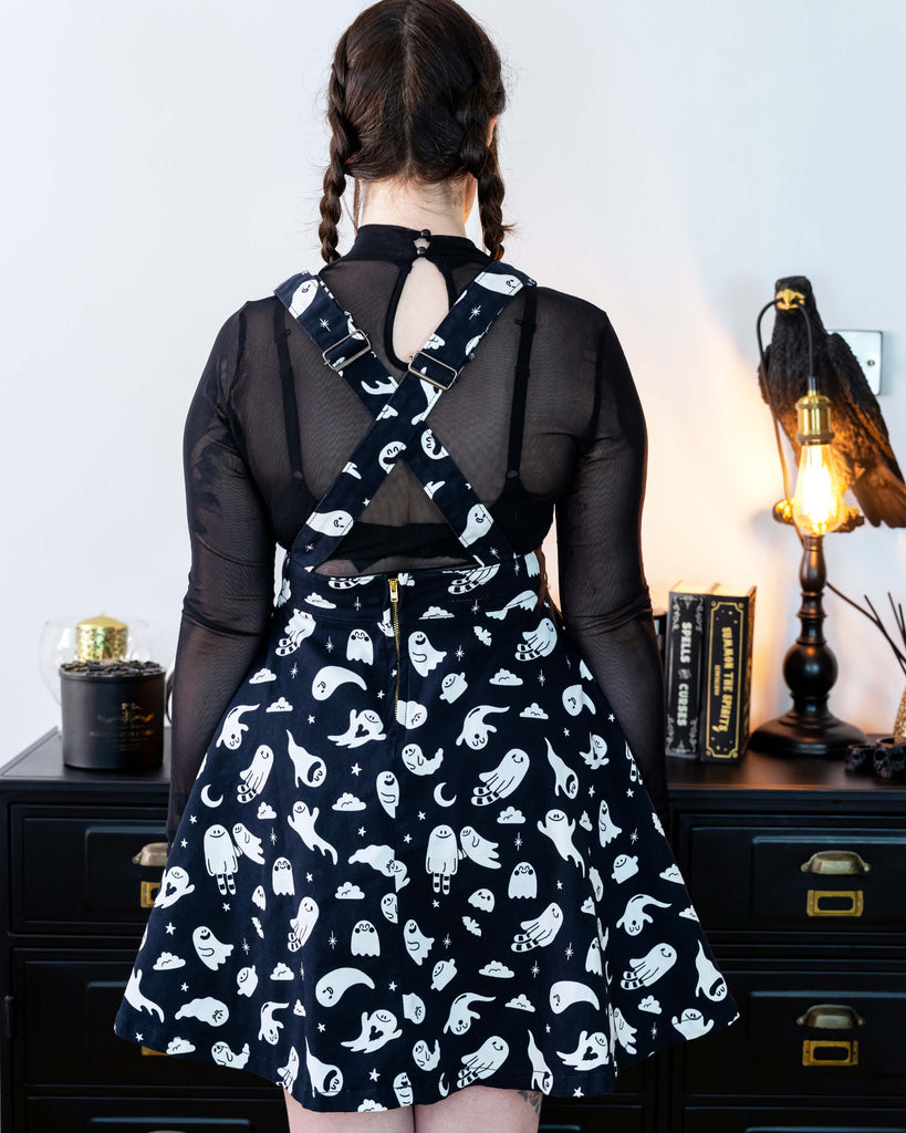 Close up of a Femme model with brown plaited hair facing away wearing a black flared pinafore dress with a ghost pattern and a mesh black top underneath in a gothic styled room with a crow lamp and black drawers.