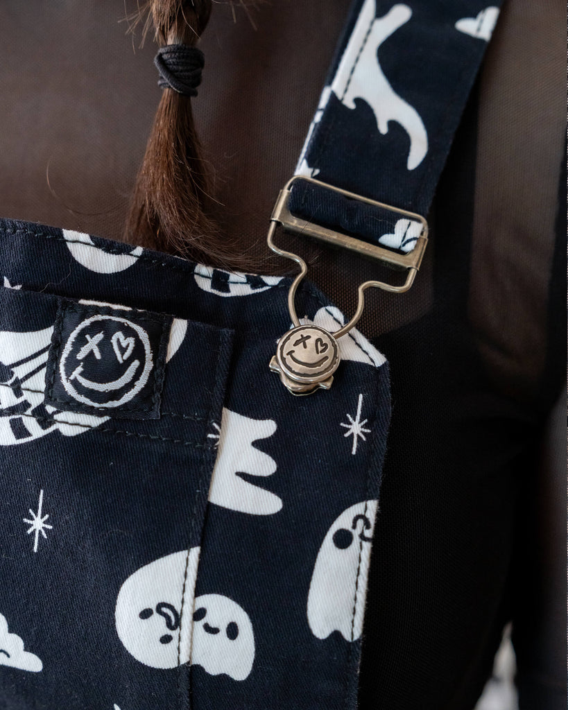 Super close up of the ghosties flared pinafore dress button buckle and rebel romance tag label detailing, a smiling face with a cross and heart for eyes. 
