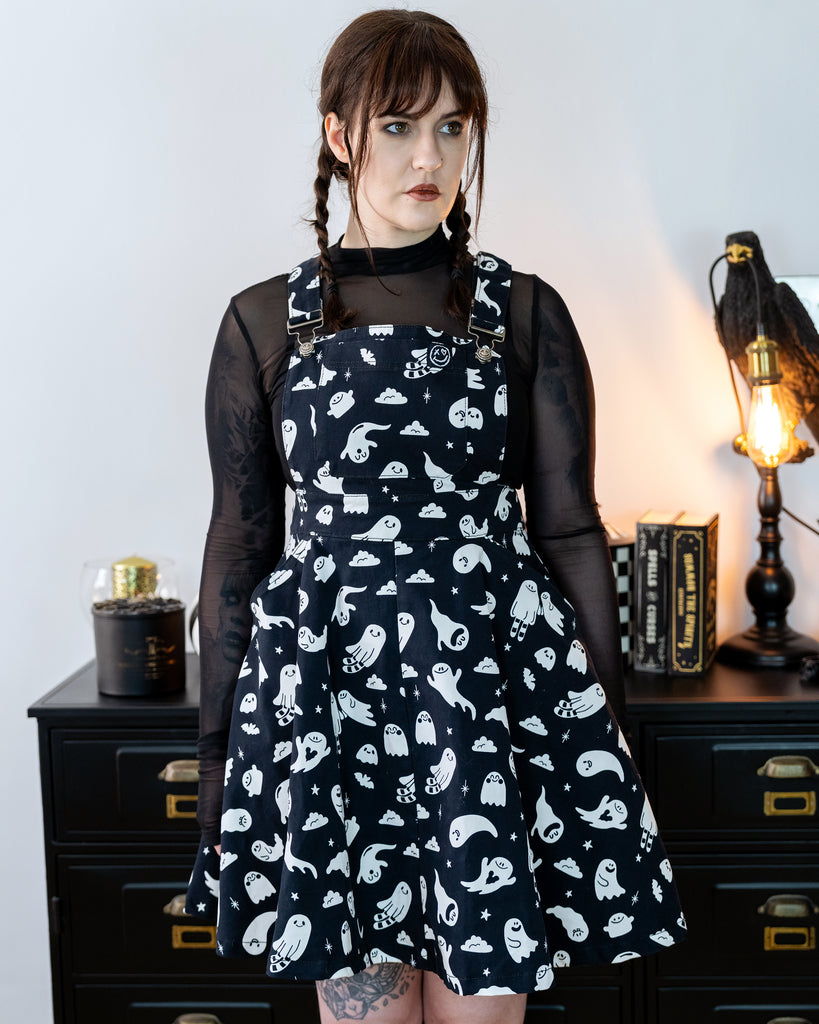 Femme model with brown plaited hair wearing a black flared pinafore dress with a ghost pattern and a mesh black top underneath in a gothic styled room with a crow lamp and black drawers.