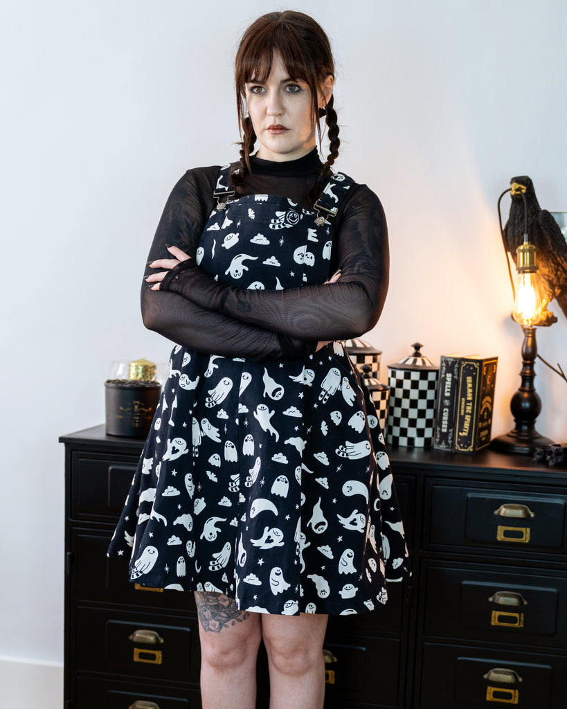Femme model with brown plaited hair wearing a black flared pinafore dress with a ghost pattern and a mesh black top underneath in a gothic styled room with a crow lamp and black drawers.