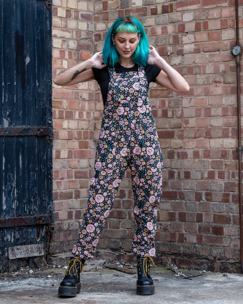 Claudia modelling Rebel Romance Eye Spy Floral Stretch Dungarees against a rustic brick wall, styled with black platform boots and black tee.