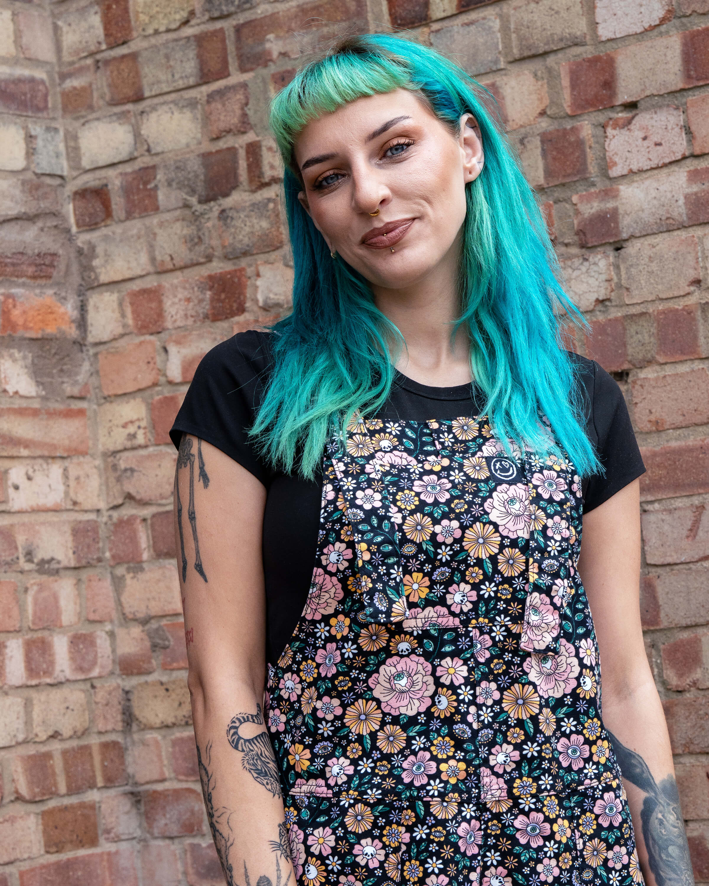 Claudia poses in the Eye Spy Floral Stretch Twill Dungarees with bold florals, skulls, and eyes, styled with a black top against a brick wall.