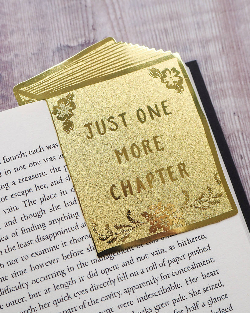 A Literary Emporium gold brass bookmark with floral designs and the phrase “Just One More Chapter,” styled on the edge of an open book. Available from Rebel Romance, a UK business celebrating alternative aesthetics and unique gifts.