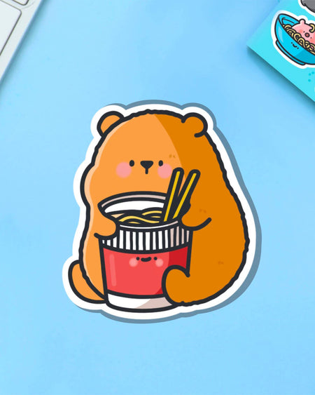 Cartoon bear eating cup noodles sticker with a happy expression, perfect for adding personality to notebooks, seen on a pastel blue surface with matching stationery.