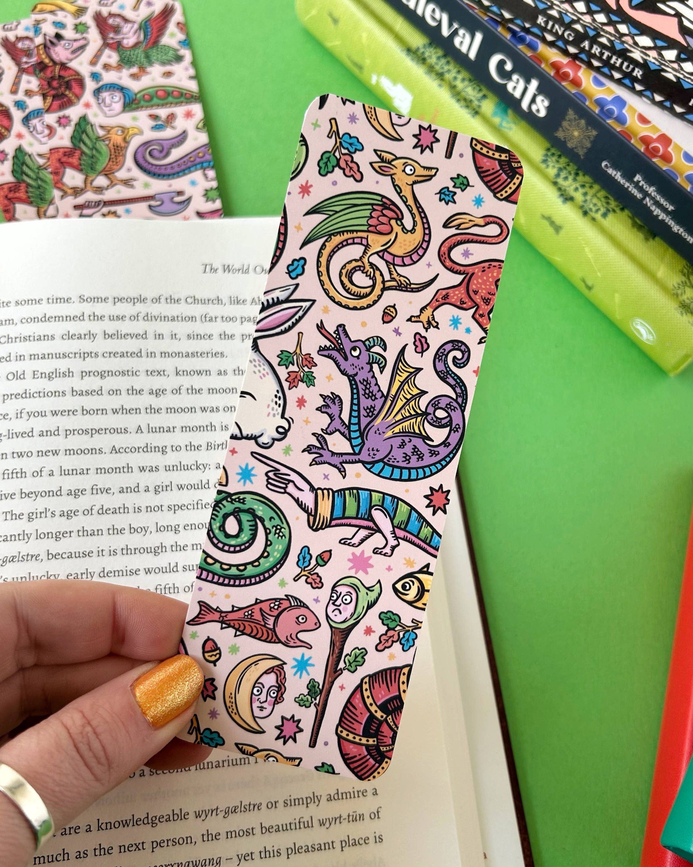 A pastel pink bookmark covered in medieval-style dragons, fish, moons, and other quirky creatures held against a green background and open book. Sold by Rebel Romance, a UK indie brand for alternative aesthetics and curious minds.