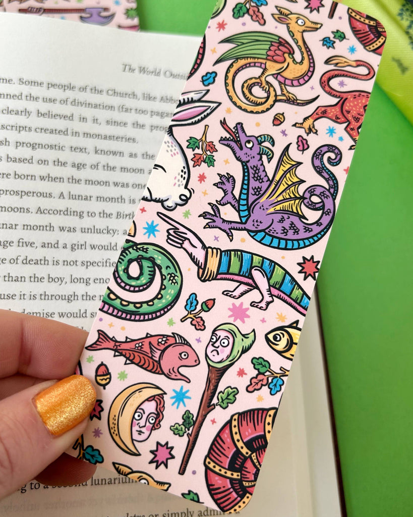 A magical illustrated bookmark filled with vibrant medieval creatures and oddities including a purple dragon, fish, moons, and leaves, shown mid-read inside an open book. Created by Stacey McEvoy Caunt, available at Rebel Romance.